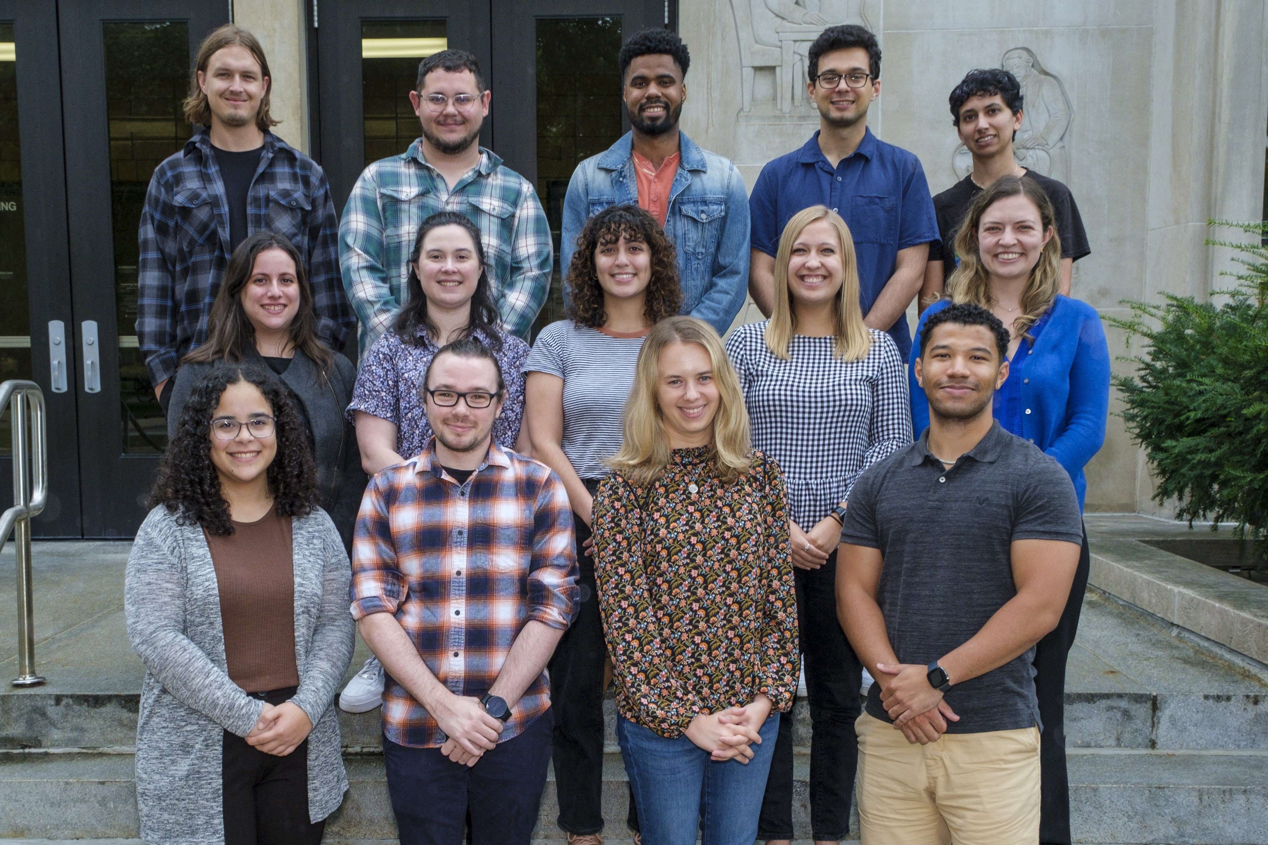 A group of graduate students smile for the camera while on the front steps of the Psychology building.