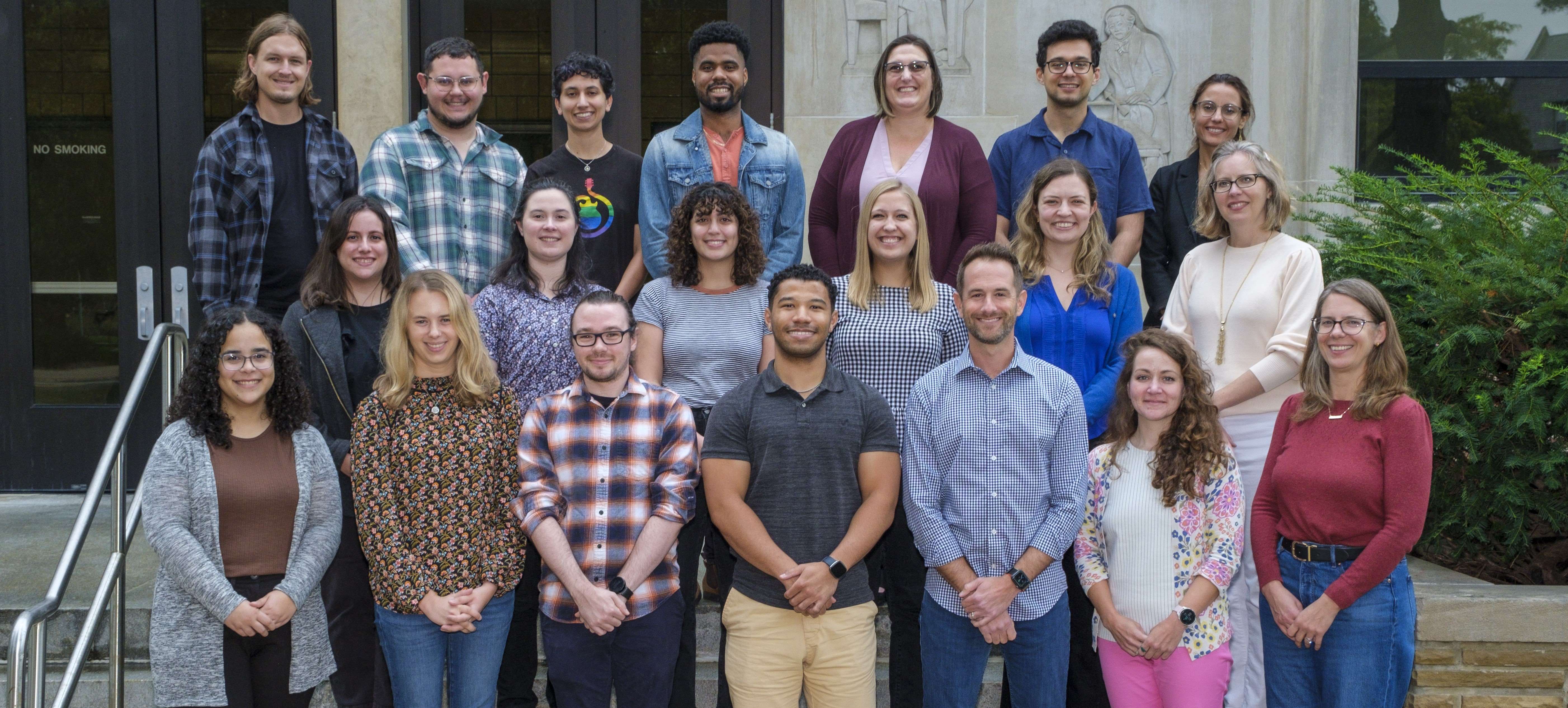 A group of faculty and graduate students smile for the camera while on the front steps of the Psychology building.