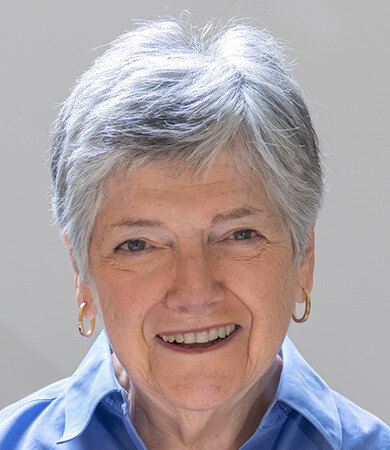 Rose Zacks smiles at the camera while wearing gold hoops earrings and a blue colored shirt. Her gray hair is short.