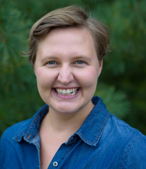 A close up headshot of Shelly DeJong. She wars a blue shirt with a green backdrop behind her.