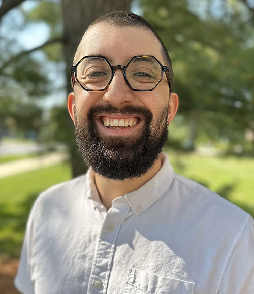 Ted Schwaba wears a light shirt and glasses and smiles directly at the camera. Behind him is a green tree and grass. 