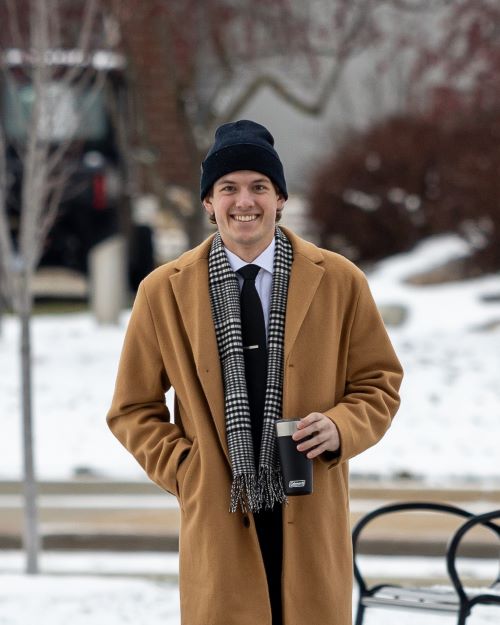 Dolan Gilbert stands outside wearing a coat and a hat and smiles at the camera.