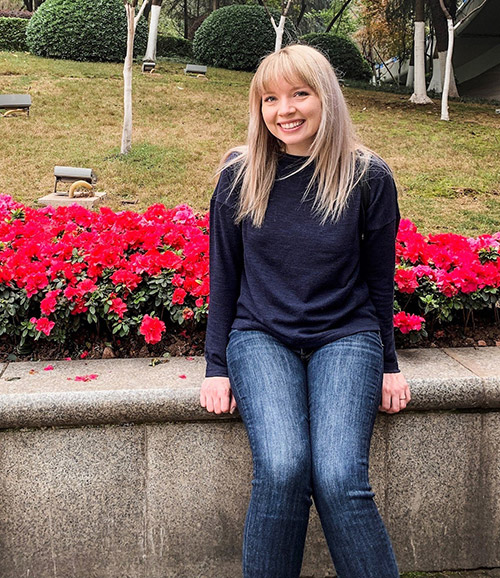 Lindsay sits on a ledge in front of red flowers. She wears a dark shirt and jeans.