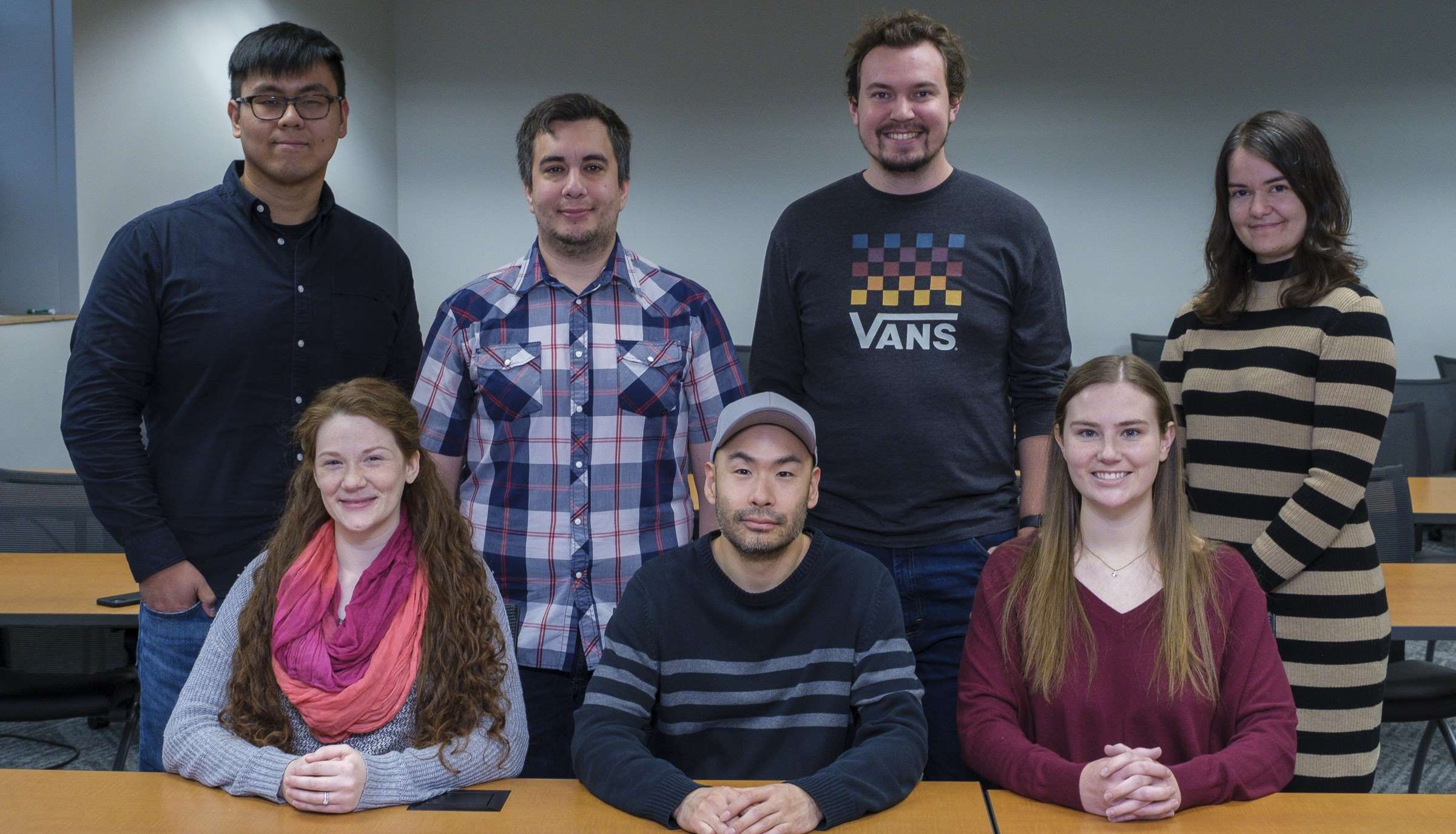 The seven current graduate students in the Cognition & Cognitive Neuroscience graduate program face the camera and smile. They are in a conference room and three sit at the tables and four stand directly behind them.