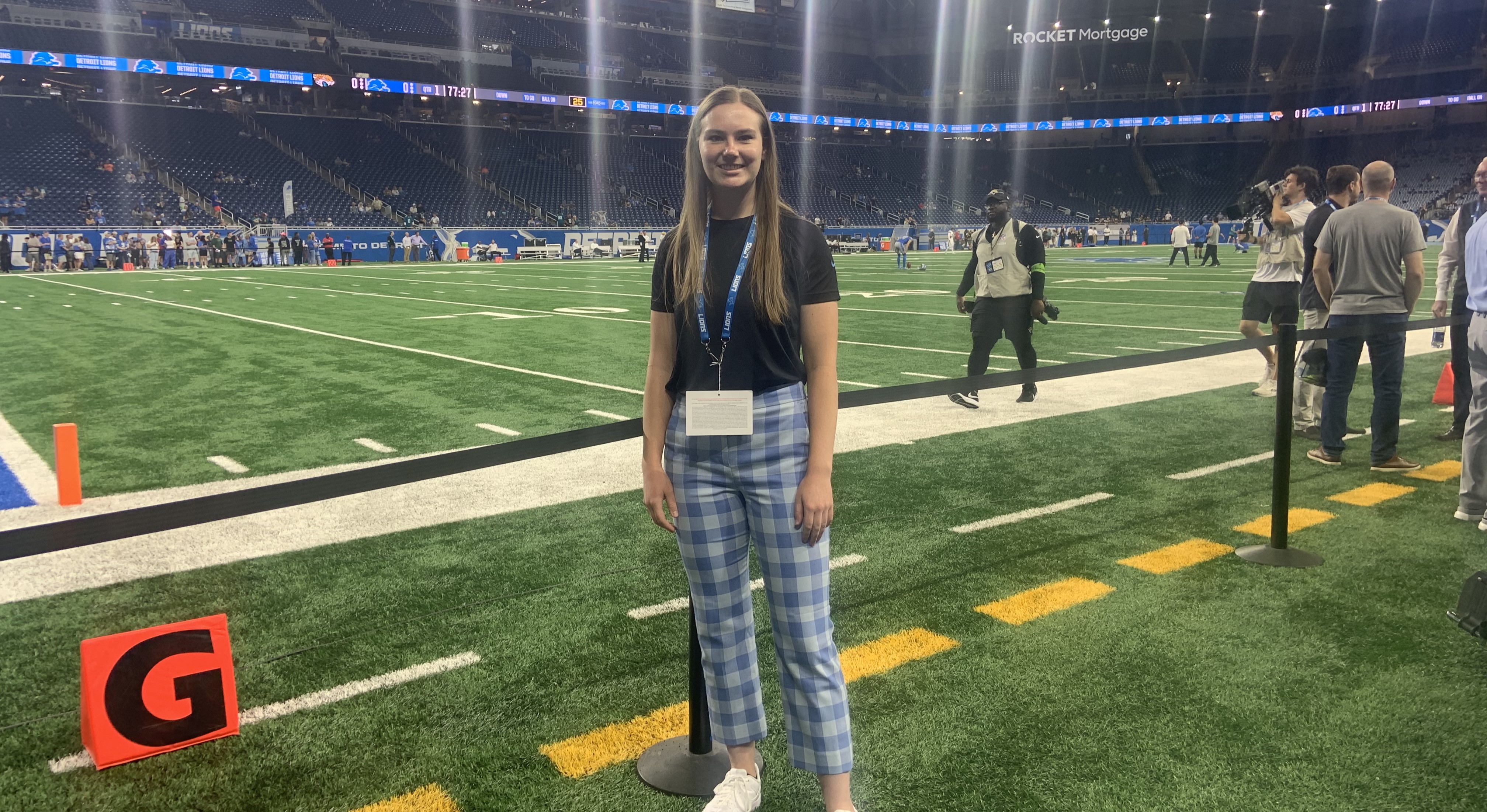 Erin Neaton wearing blue plaid pants, a dark shirt, and a lanyard stands on the sidelines of the Detroit Lions football field before a game..