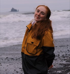 Shannon Gielow stands on the shoreline with water behind her. She smiles at the camera while wearing a tan and black jacket.