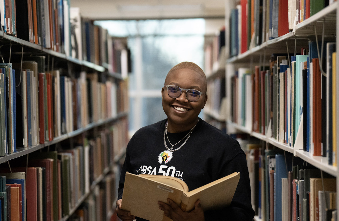 Joya Bailey holds a book open at the library and smiles at the camera.